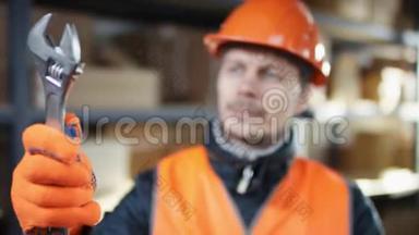 与面包和胡子工人一起工作，戴着头盔和工作服，站在架子旁边，检查一个可调节的扳手。